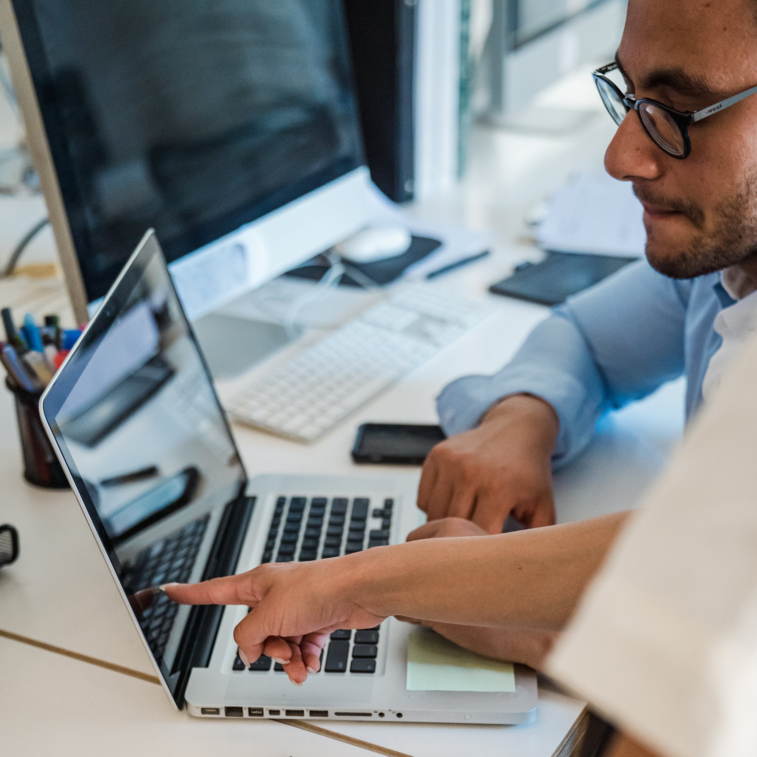 One person sits in front of a laptop at a desk while the hand of a second person points at the laptop screen